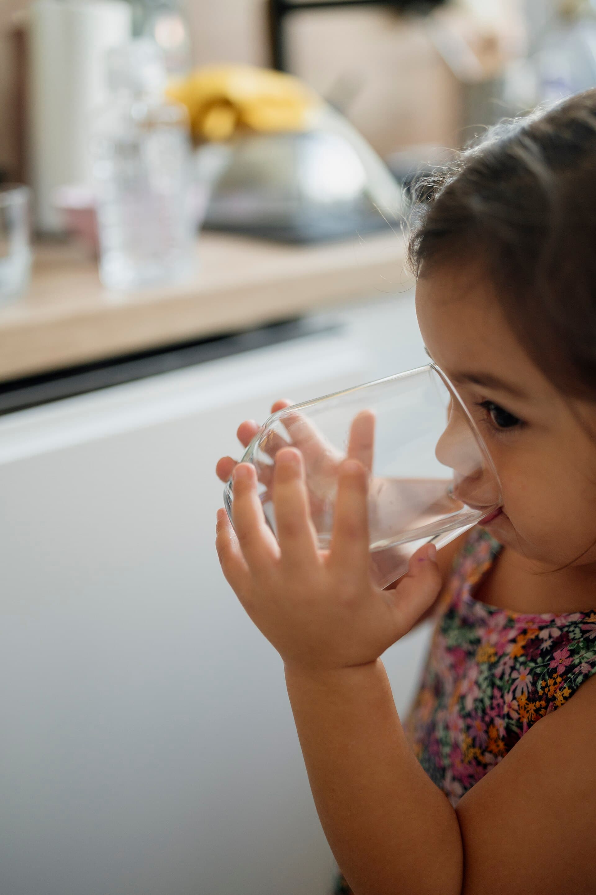 A young child drinks from a glass while standing in a kitchen